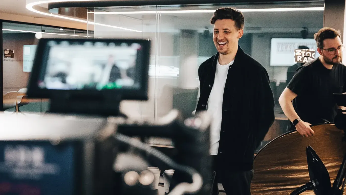 Mark G Smith laughs between takes, seen past the camera monitor, with Lewis Rees on the right